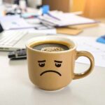 Taza de café con carita triste sobre un escritorio, ilustrando malestar o efectos negativos del exceso de café
